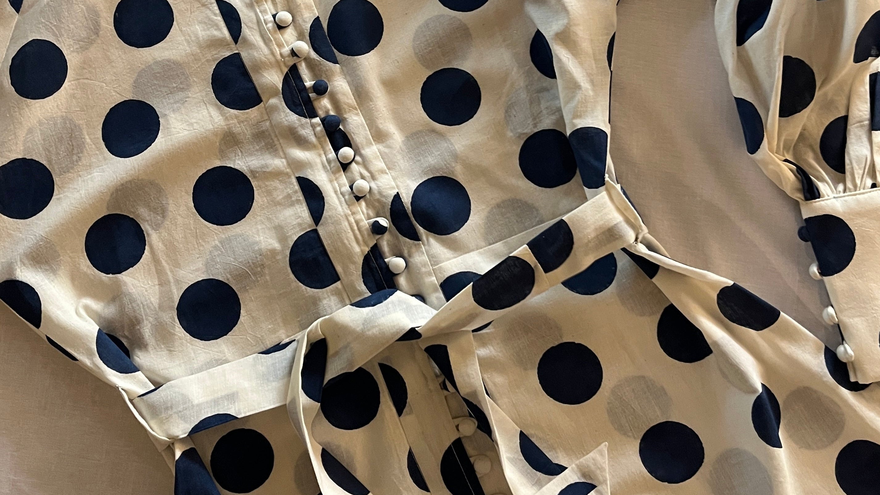 A cream-colored long-sleeve blouse with a navy blue and gray polka dot pattern, featuring a button-up front and a matching tie belt at the waist, laid flat on a light-colored fabric background.