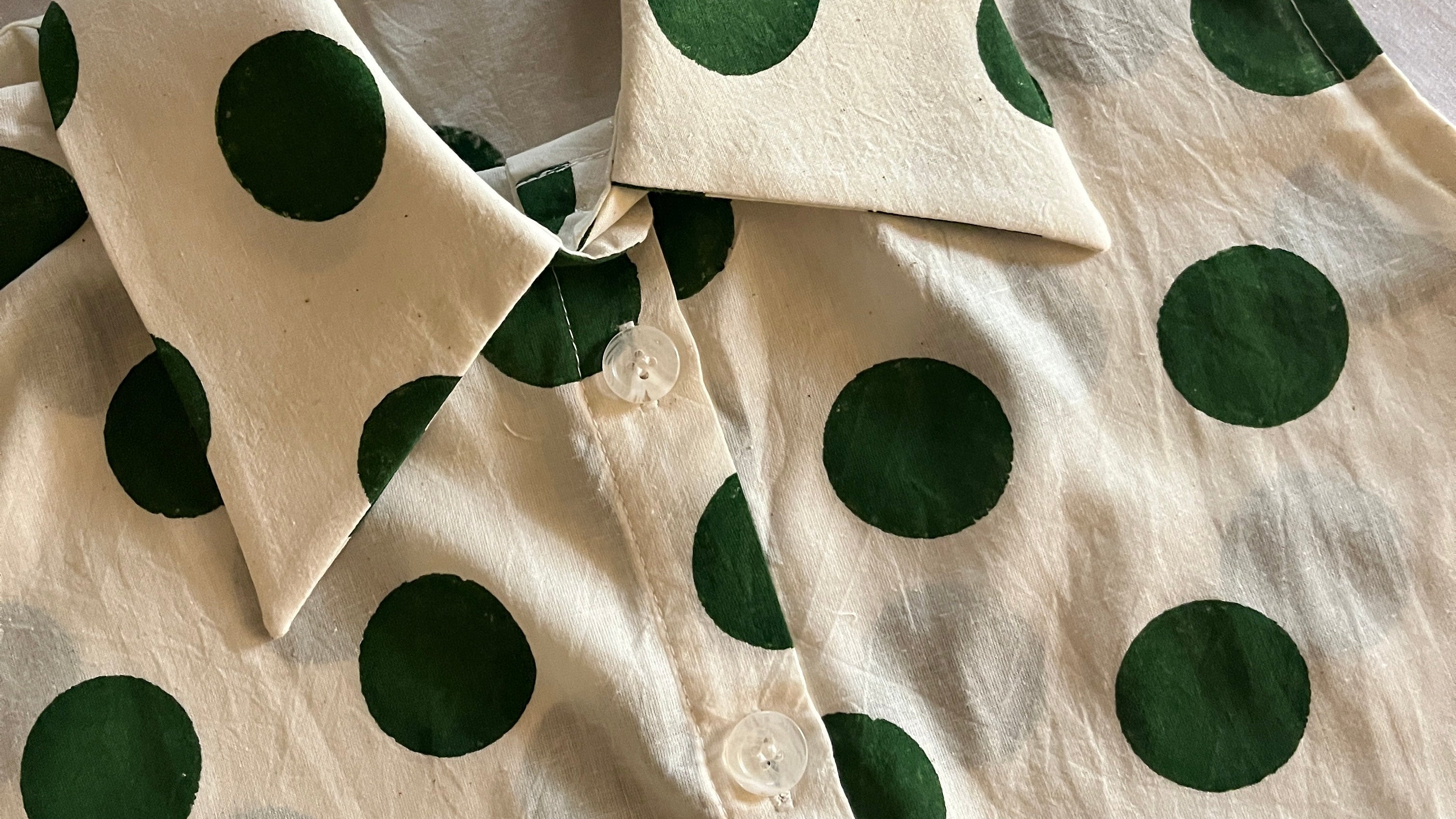 A close-up of a cream-colored button-up shirt with a dark green polka dot pattern, laid flat on a white fabric background.