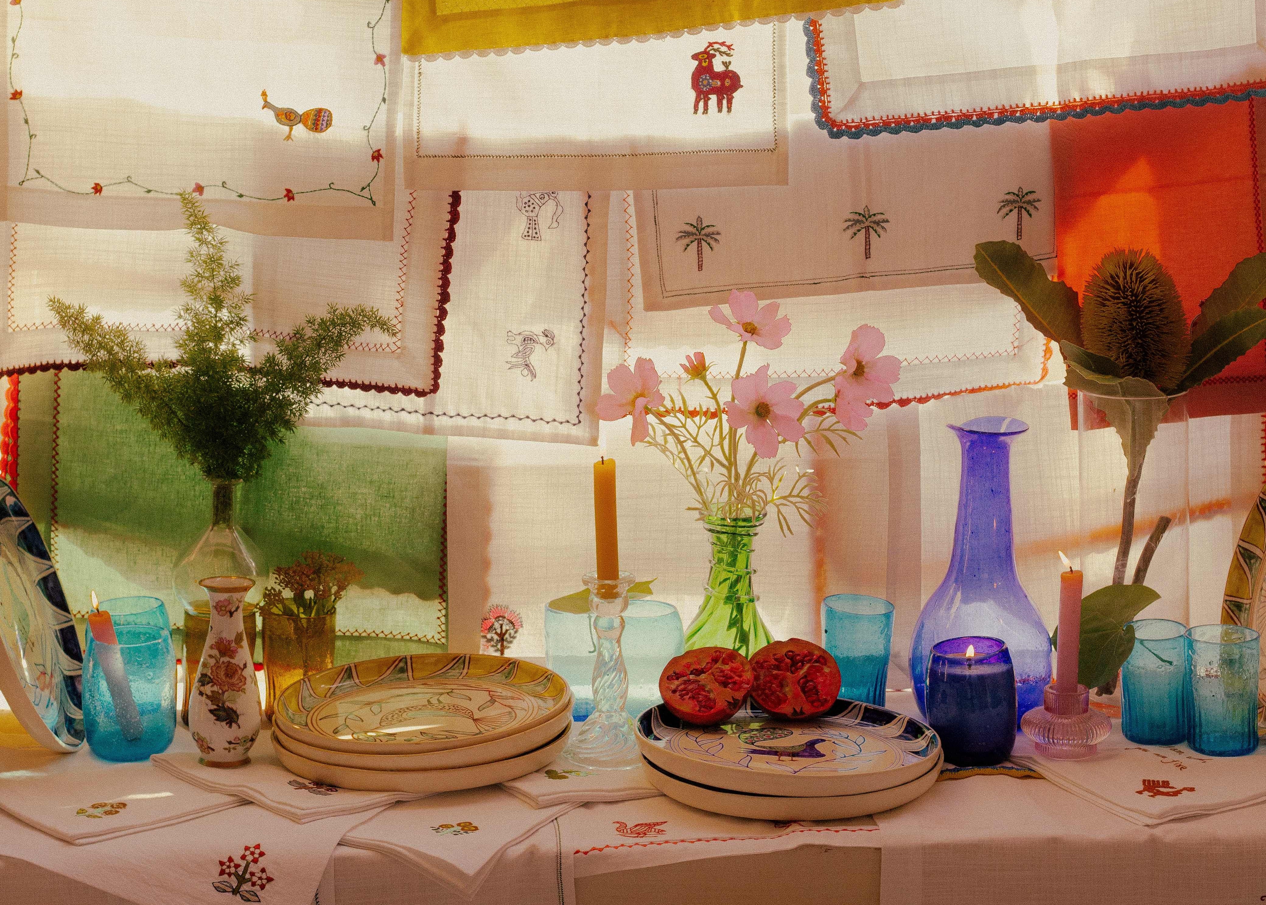 A styled tabletop display featuring embroidered table linens, colorful glassware in blue and green, candles, plates, and dried botanicals, with embroidered fabric panels hanging in the background.