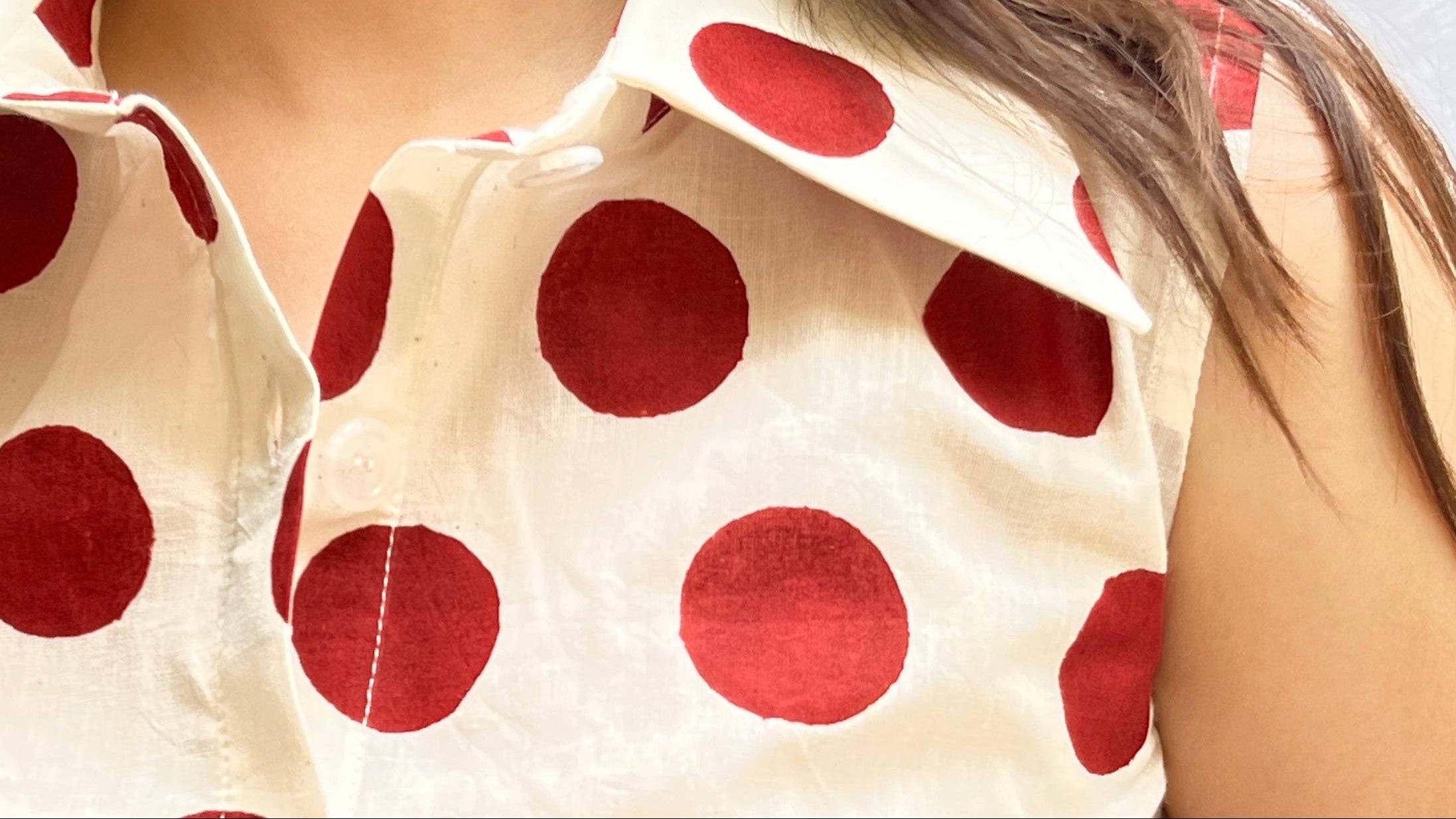 A close-up of a woman wearing a sleeveless collared button-front top with a red polka dot pattern on a cream background, against a light textured backdrop.